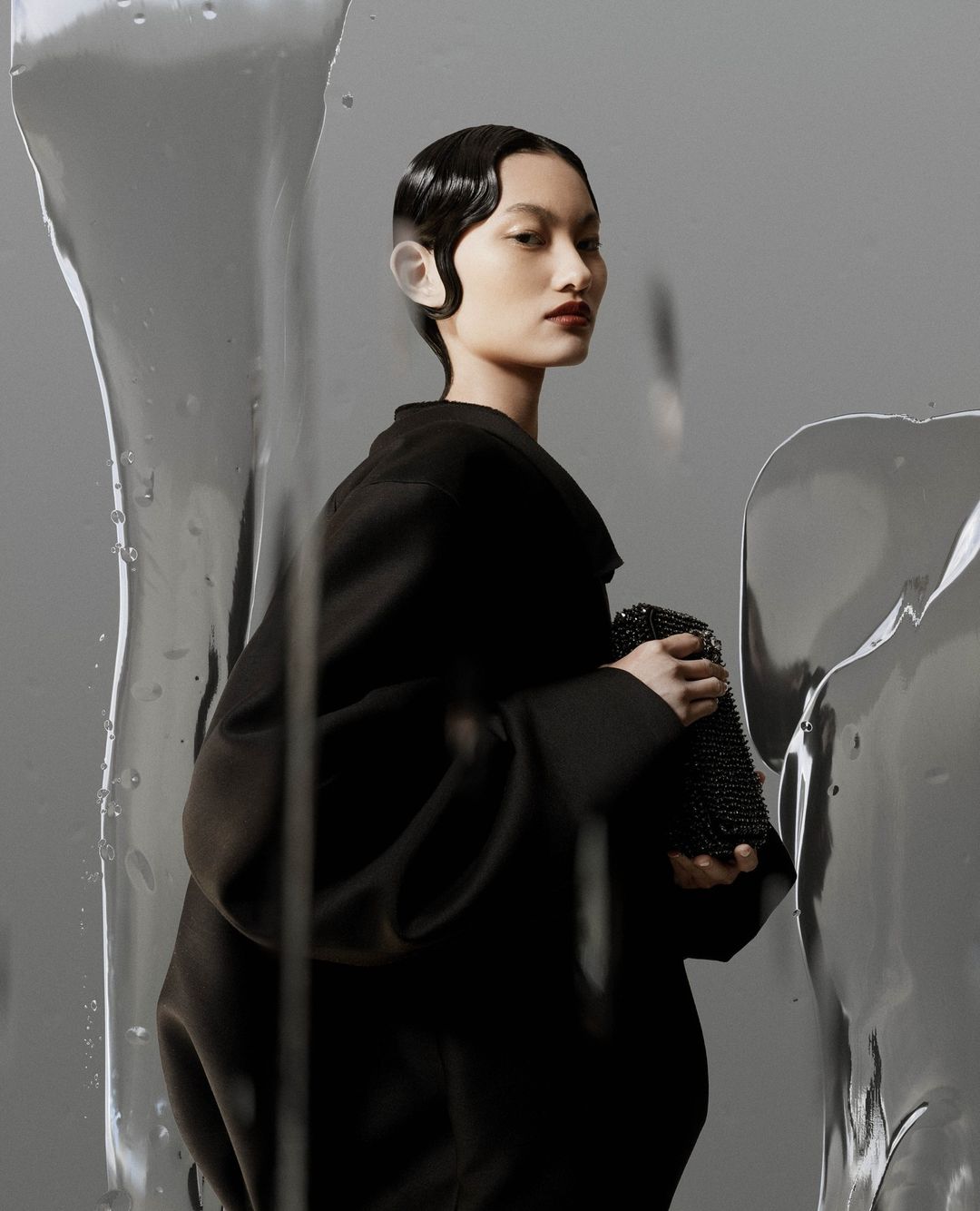Elegant Asian woman in black coat holding beaded purse, clean makeup, and wet-look hair styled in waves, studio shot on grey backdrop.