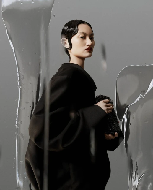 Elegant Asian woman in black coat holding beaded purse, clean makeup, and wet-look hair styled in waves, studio shot on grey backdrop.