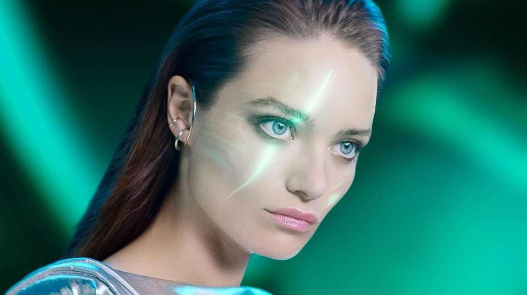 Close-up beauty shot of an adult woman with light skin, light eyes, and dark hair, wearing modern ear jewelry against a green background.