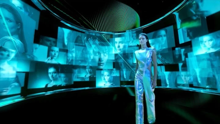 Woman in a metallic jumpsuit poses in a futuristic digital environment with glowing blue screens displaying various portraits.