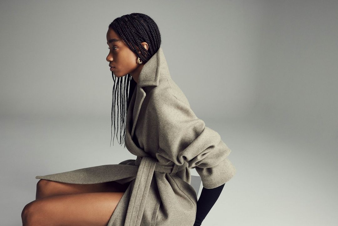 Woman with braided hair sits in a grey wrap coat in a studio with a seamless background in a medium shot.
