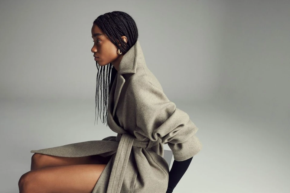 Woman with braided hair sits in a grey wrap coat in a studio with a seamless background in a medium shot.