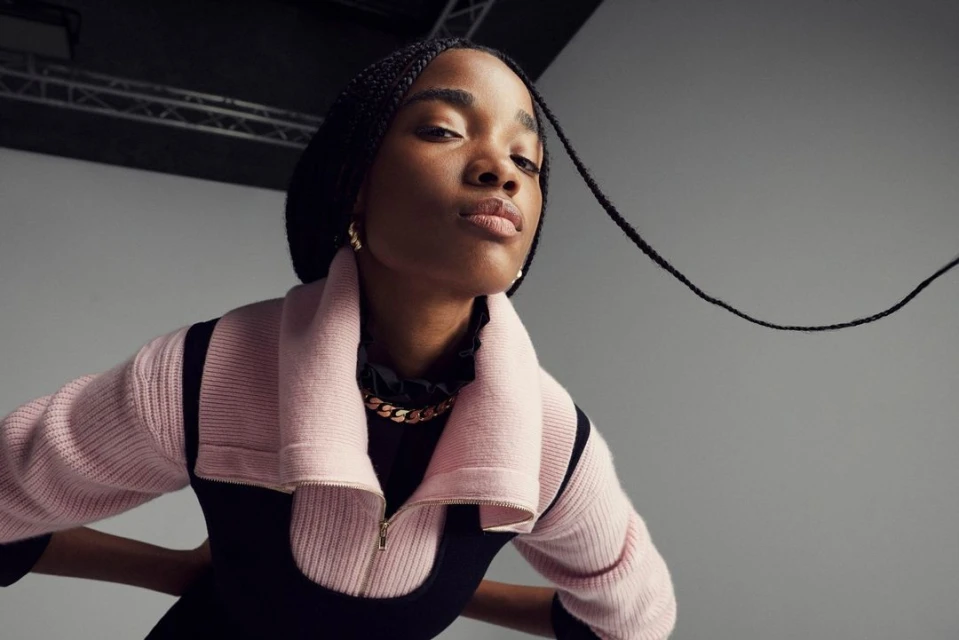 A dark-skinned adult female is photographed in a studio from a low angle wearing a pink jacket, gold chain, and hoop earrings.