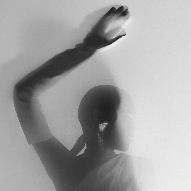 Monochrome artistic photograph of a person with an upraised arm and hand, draped cloth, earrings, and a soft blurred aesthetic.