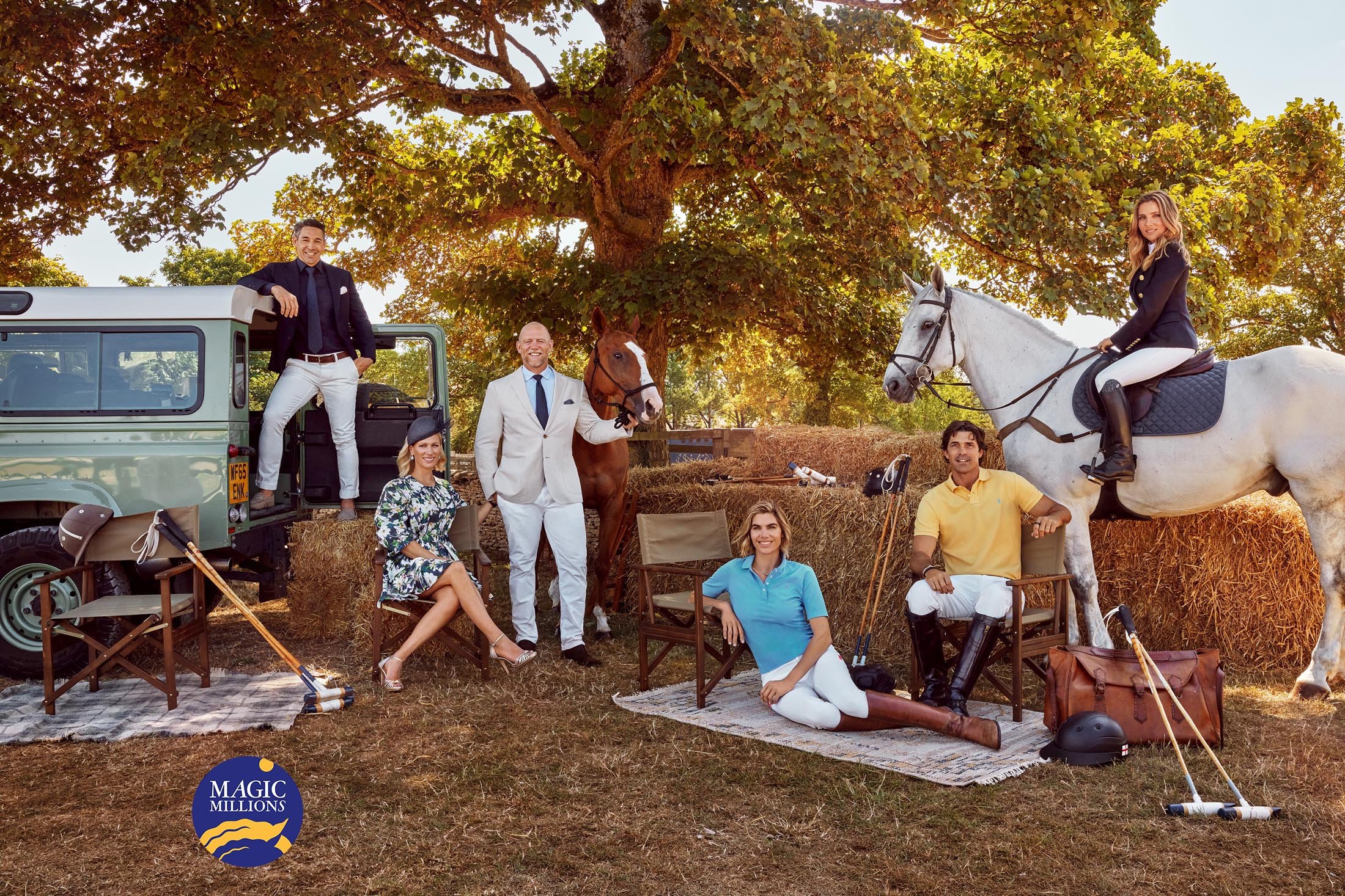 Group of people and horses gathered in a field with a Land Rover, hay bales, and polo equipment under a large tree on a sunny day.