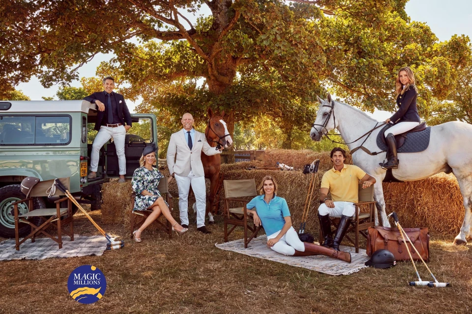 Group of people and horses gathered in a field with a Land Rover, hay bales, and polo equipment under a large tree on a sunny day.