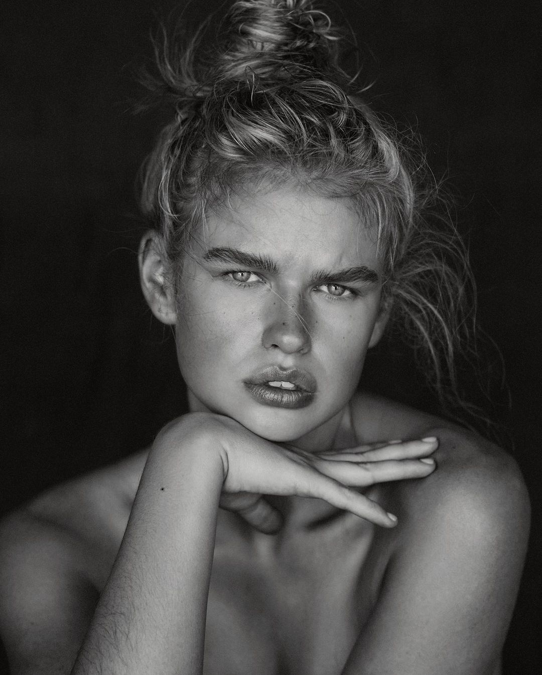 Striking black and white portrait of a woman with blonde hair in a bun, chin resting on her hand, against a dark background.