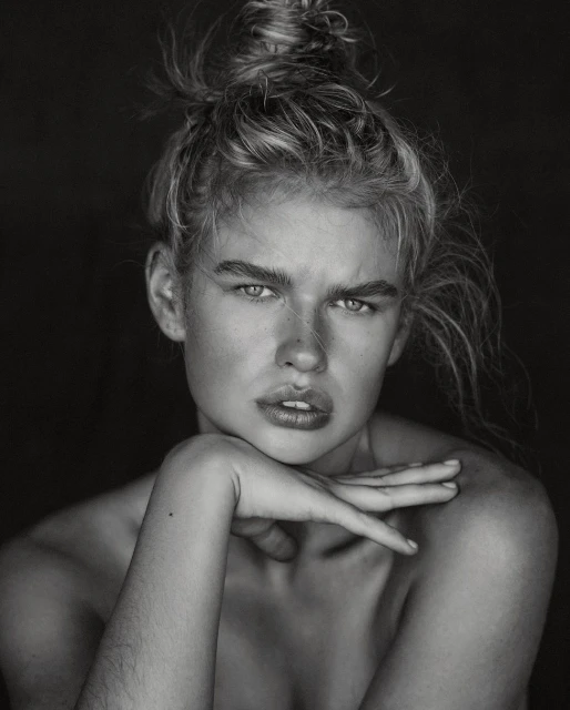Striking black and white portrait of a woman with blonde hair in a bun, chin resting on her hand, against a dark background.