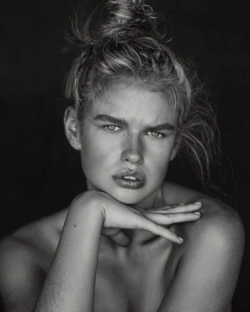 Striking black and white portrait of a woman with blonde hair in a bun, chin resting on her hand, against a dark background.