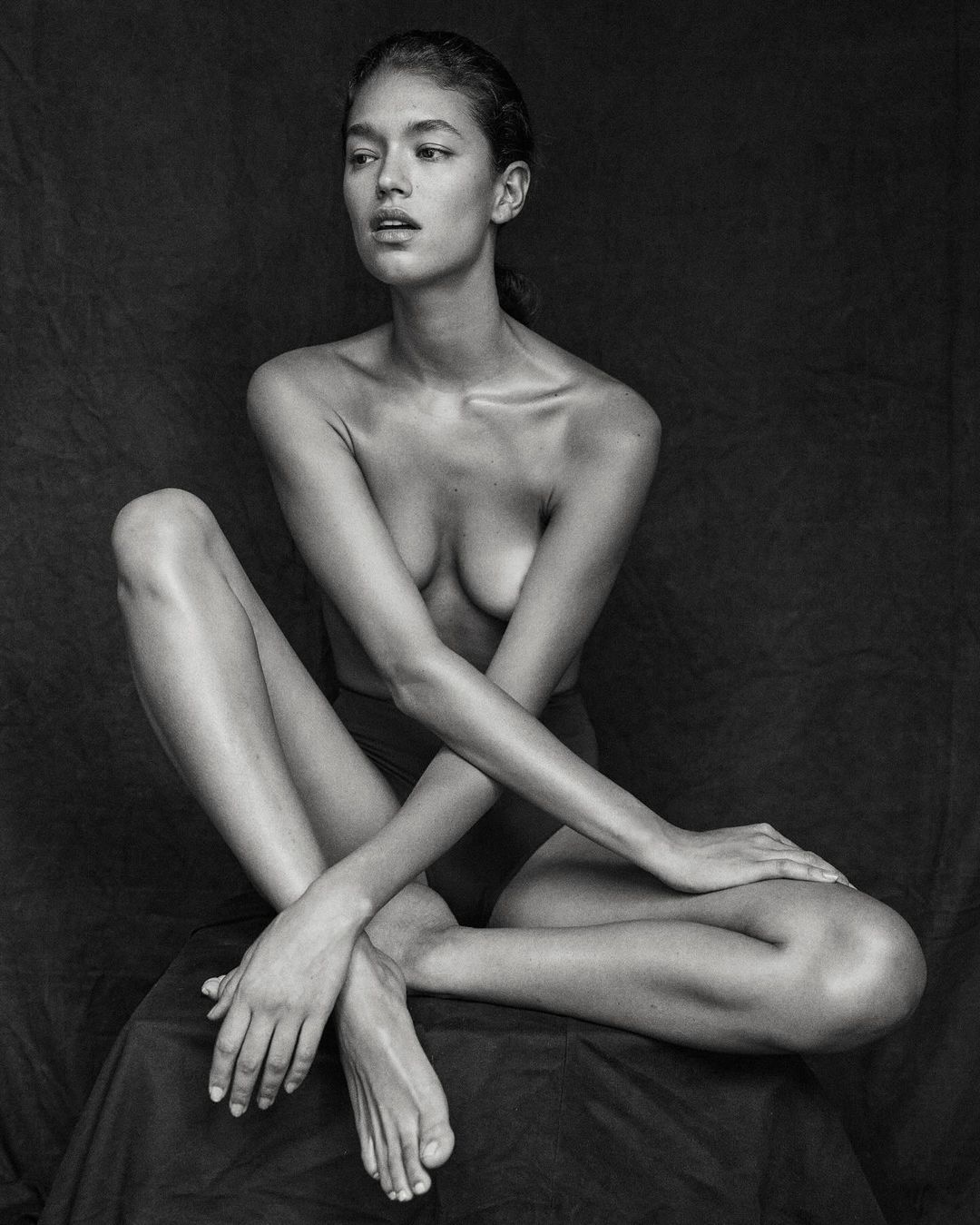 Black-and-white portrait of a bare-chested woman with crossed arms sitting on a fabric draped surface in a dimly lit indoor setting.