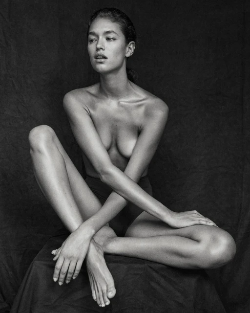 Black-and-white portrait of a bare-chested woman with crossed arms sitting on a fabric draped surface in a dimly lit indoor setting.
