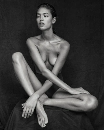 Black-and-white portrait of a bare-chested woman with crossed arms sitting on a fabric draped surface in a dimly lit indoor setting.
