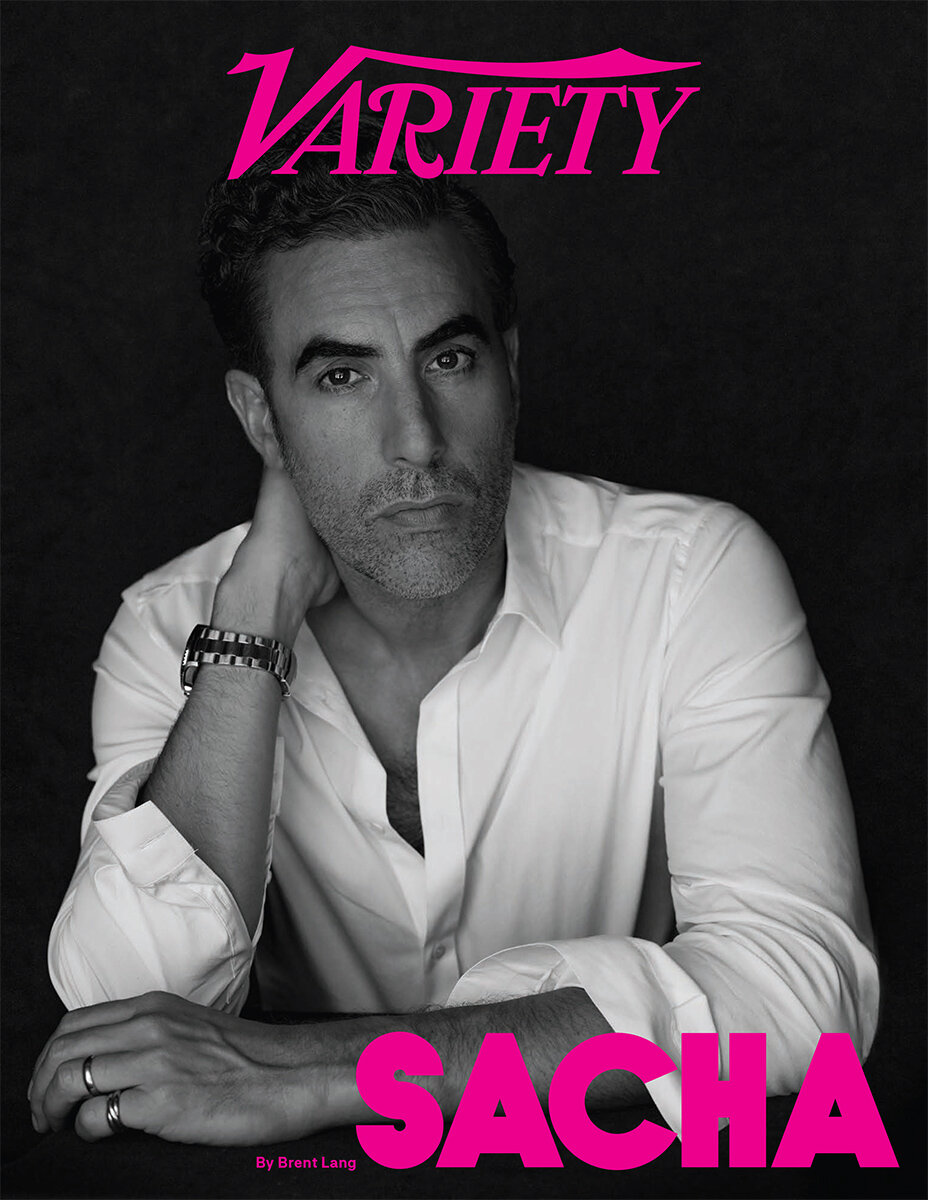 Magazine cover featuring Sacha Baron Cohen in a white collared shirt with a silver watch, set against a dark background.