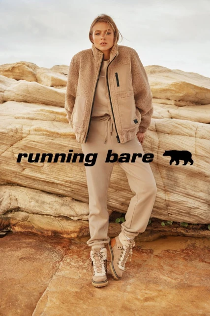 A fair-skinned woman models sportswear on layered rocks in front of a neutral sky, wearing a fleece jacket, sweatshirt, sweatpants, and boots.