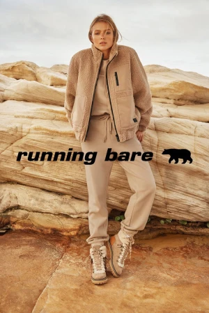 A fair-skinned woman models sportswear on layered rocks in front of a neutral sky, wearing a fleece jacket, sweatshirt, sweatpants, and boots.