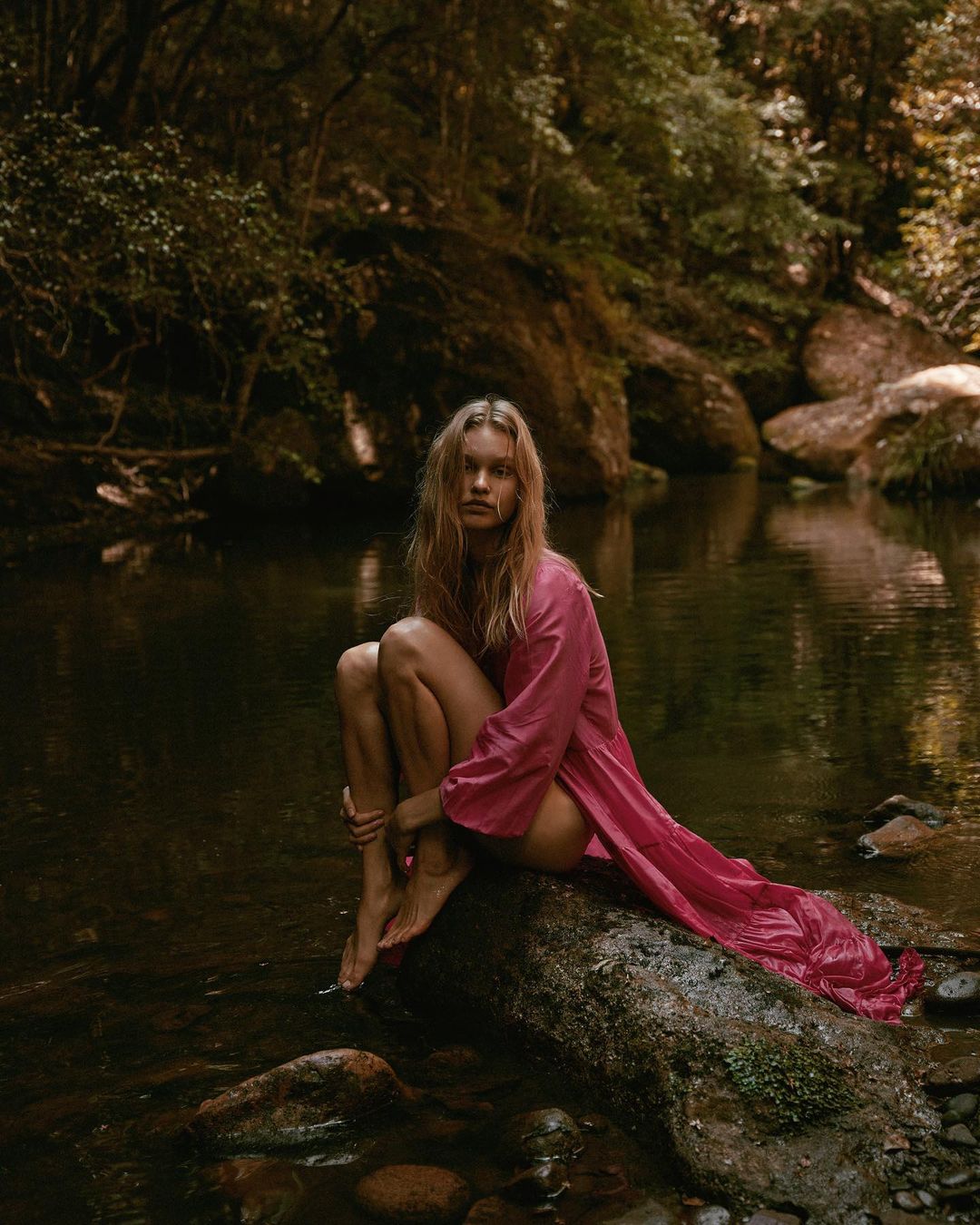 Woman with long blonde hair sitting on a rock in a stream, wearing a pink robe, surrounded by lush forest and reflective water.