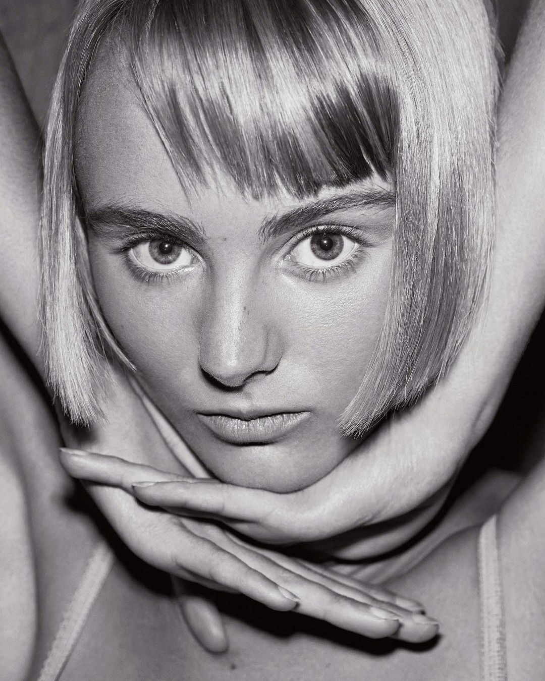 Close-up portrait of a young fair-skinned woman with blonde bob, her hands framing her face, in a monochromatic indoor setting.