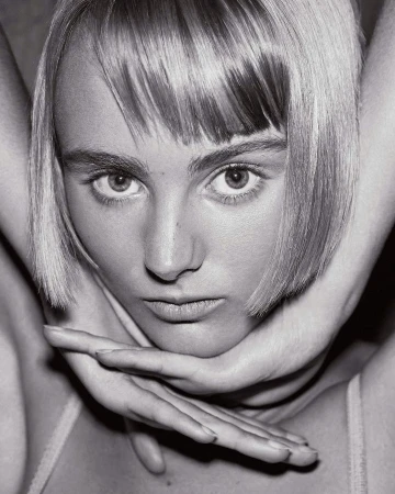 Close-up portrait of a young fair-skinned woman with blonde bob, her hands framing her face, in a monochromatic indoor setting.