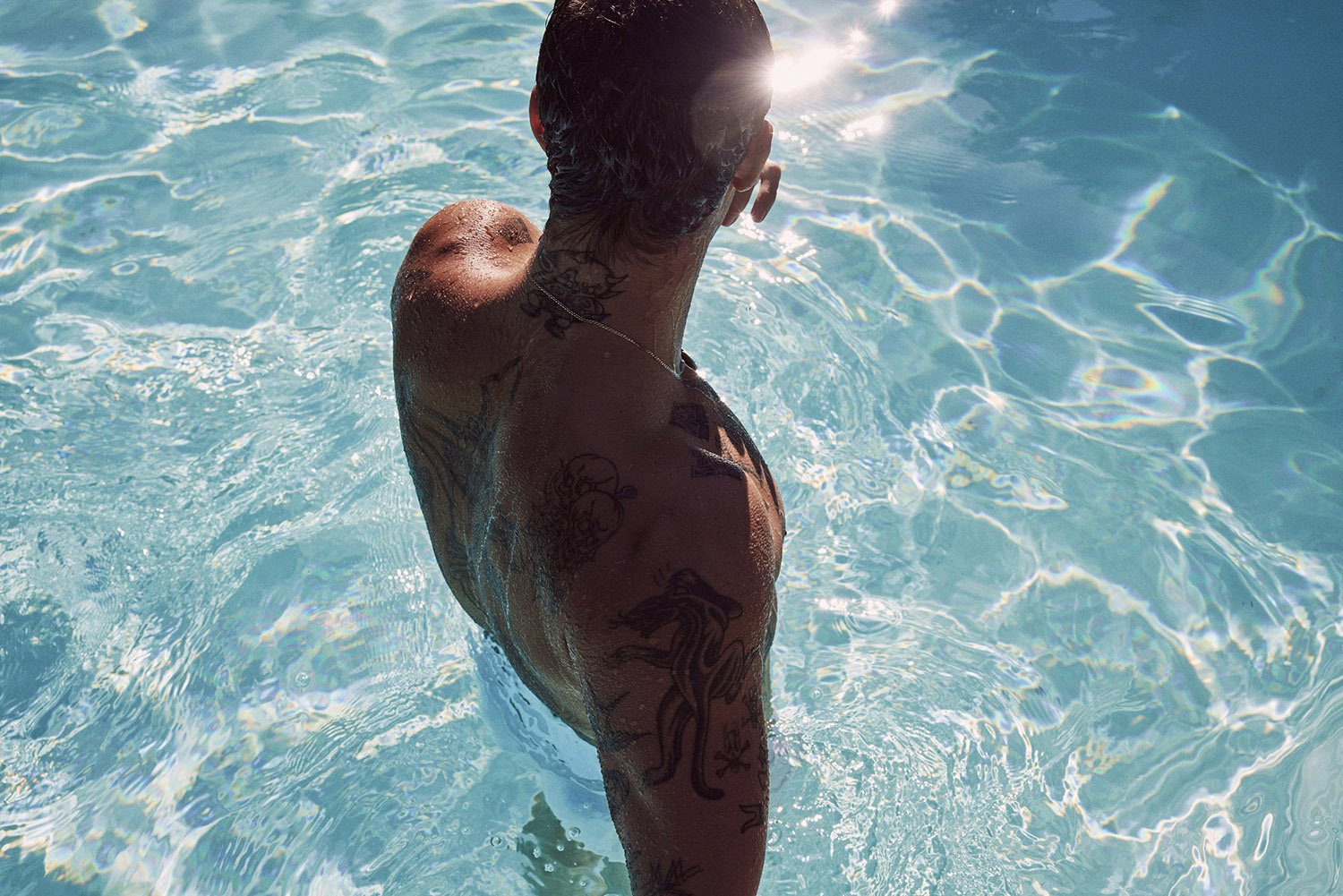 Adult male partially submerged in clear blue swimming pool water. Light glares off the ripples in the water on a sunny day.