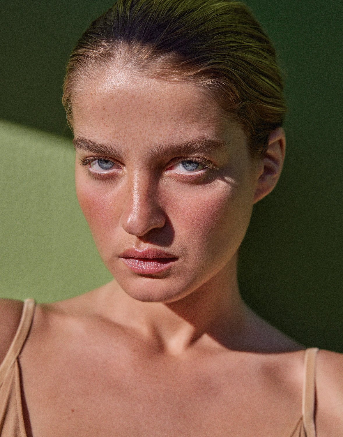 Close-up portrait of a young adult female with fair skin and blue eyes. The woman is facing forward against a green background with natural lighting.