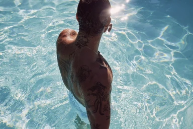 Shirtless tattooed man standing in bright turquoise swimming pool water with sunlight glinting off surface
