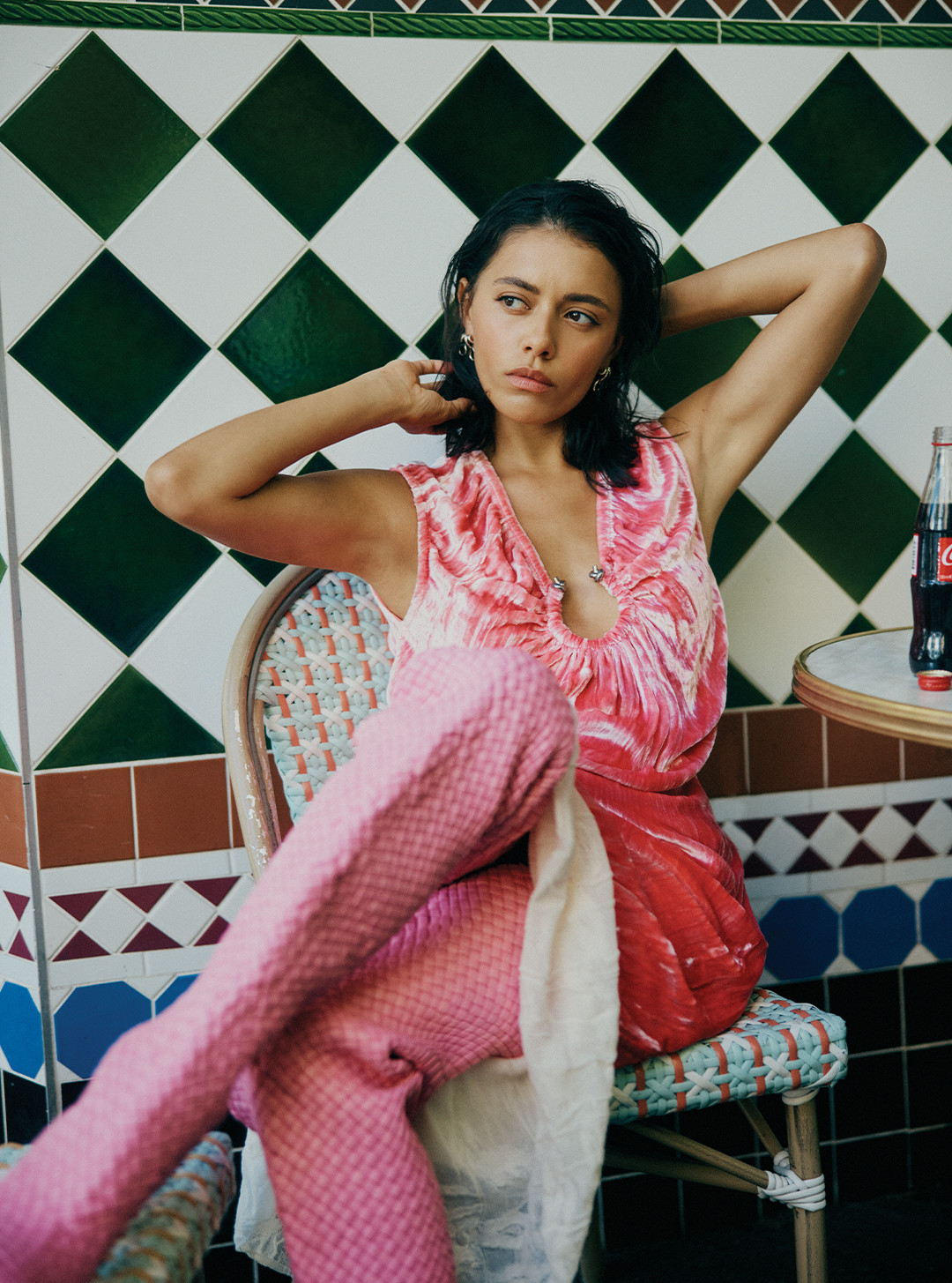 Woman with short dark hair poses in a pink top and textured pants, seated indoors with a Coca-Cola bottle on a marble-topped table.