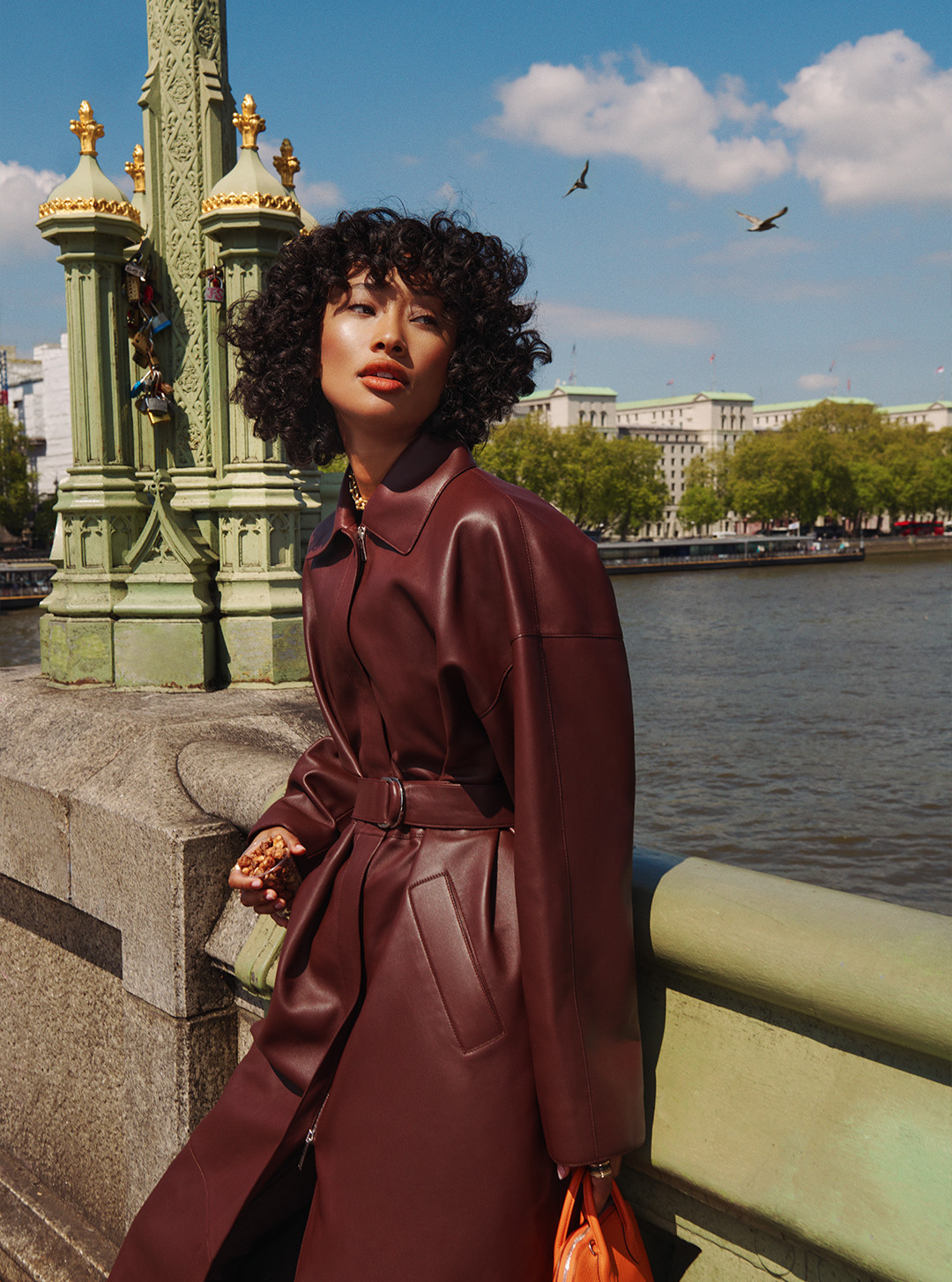 An Asian woman with curly black hair wears a wine-red leather trench coat. She is standing on a bridge with a purse in hand.