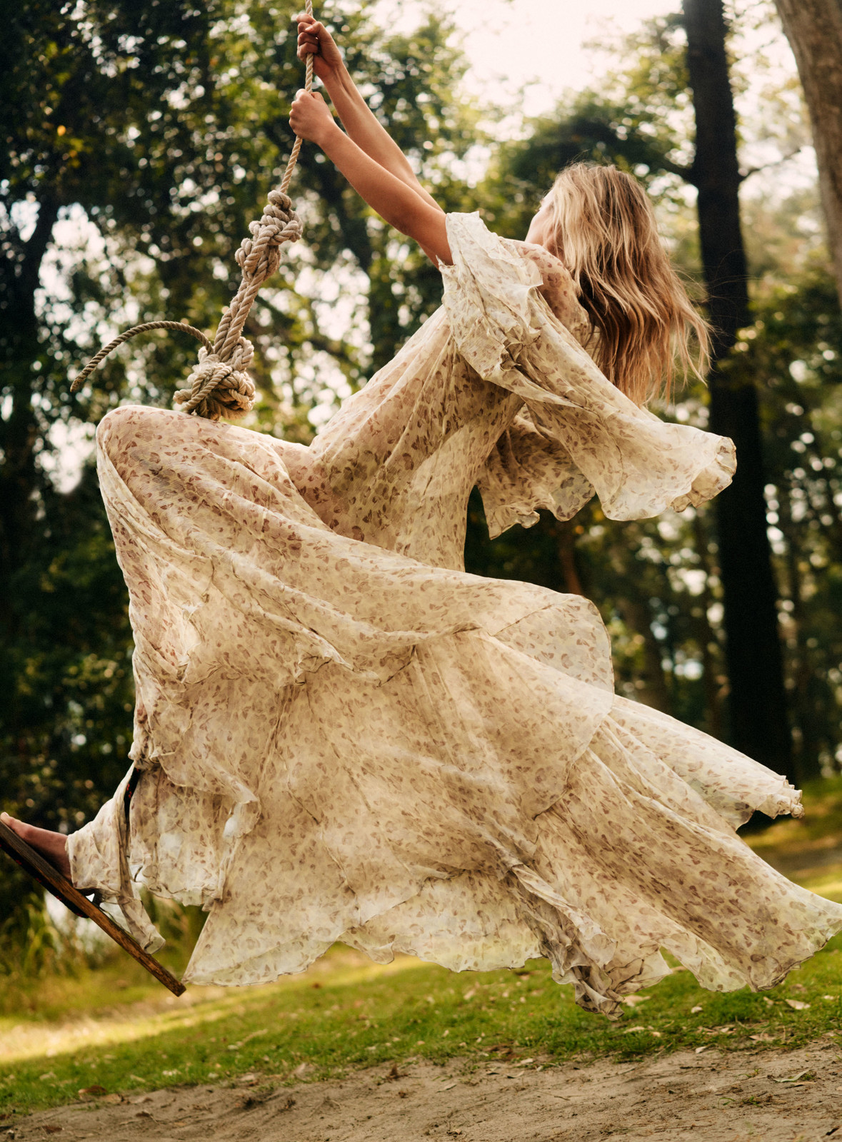 Woman in a long flowing dress swings on a rope swing hanging from a tree in a lush green forest. Summery and carefree.