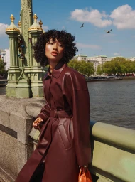 Woman with dark curly hair wearing a burgundy leather coat standing near a green bridge overlooking a river