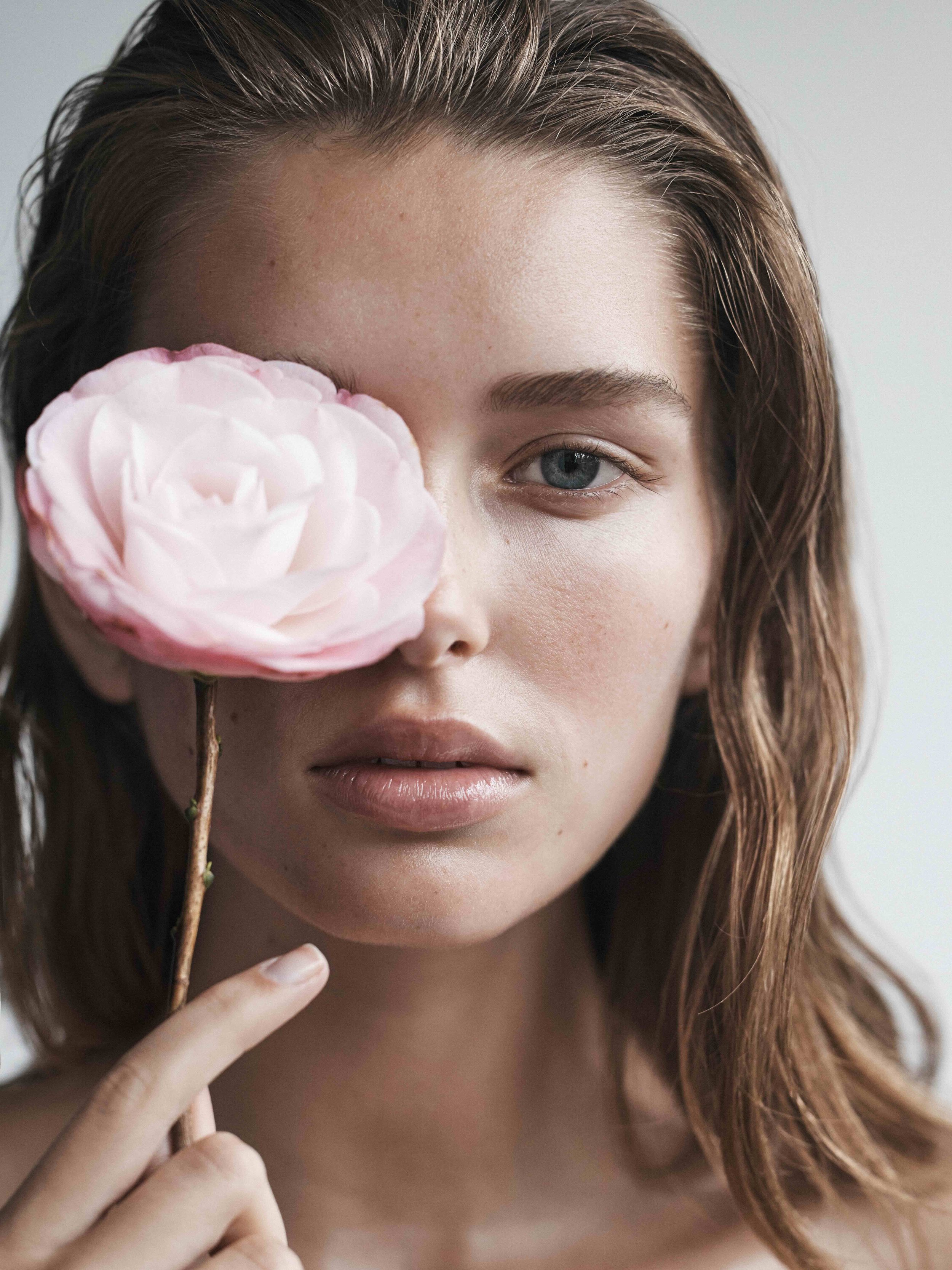 Close-up portrait of a young adult female with fair skin, brown hair, holding a light pink flower covering her left eye in a beauty shot.