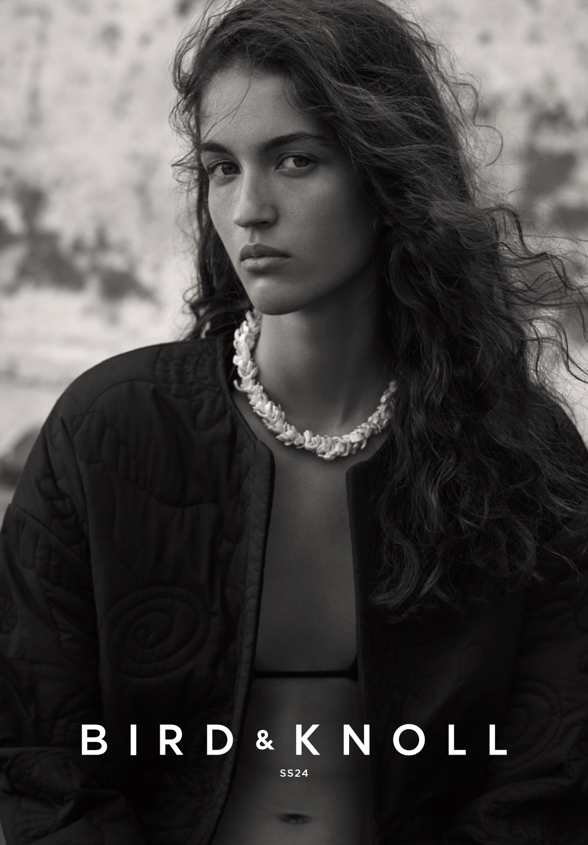 Monochrome portrait of a young adult female model with wavy hair wearing a seashell necklace and a quilted jacket outdoors.