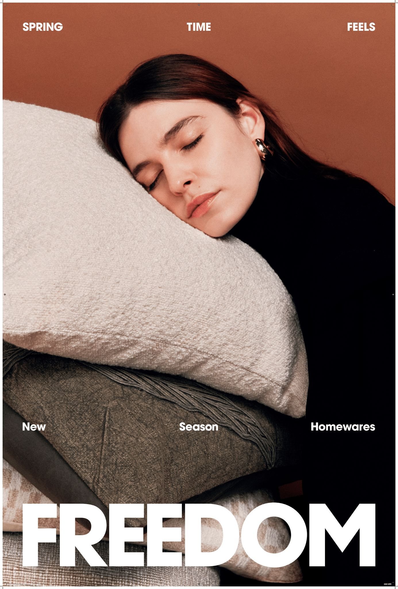 A woman with dark hair rests her face on a stack of soft pillows against a warm brown background in a cozy and inviting setting.