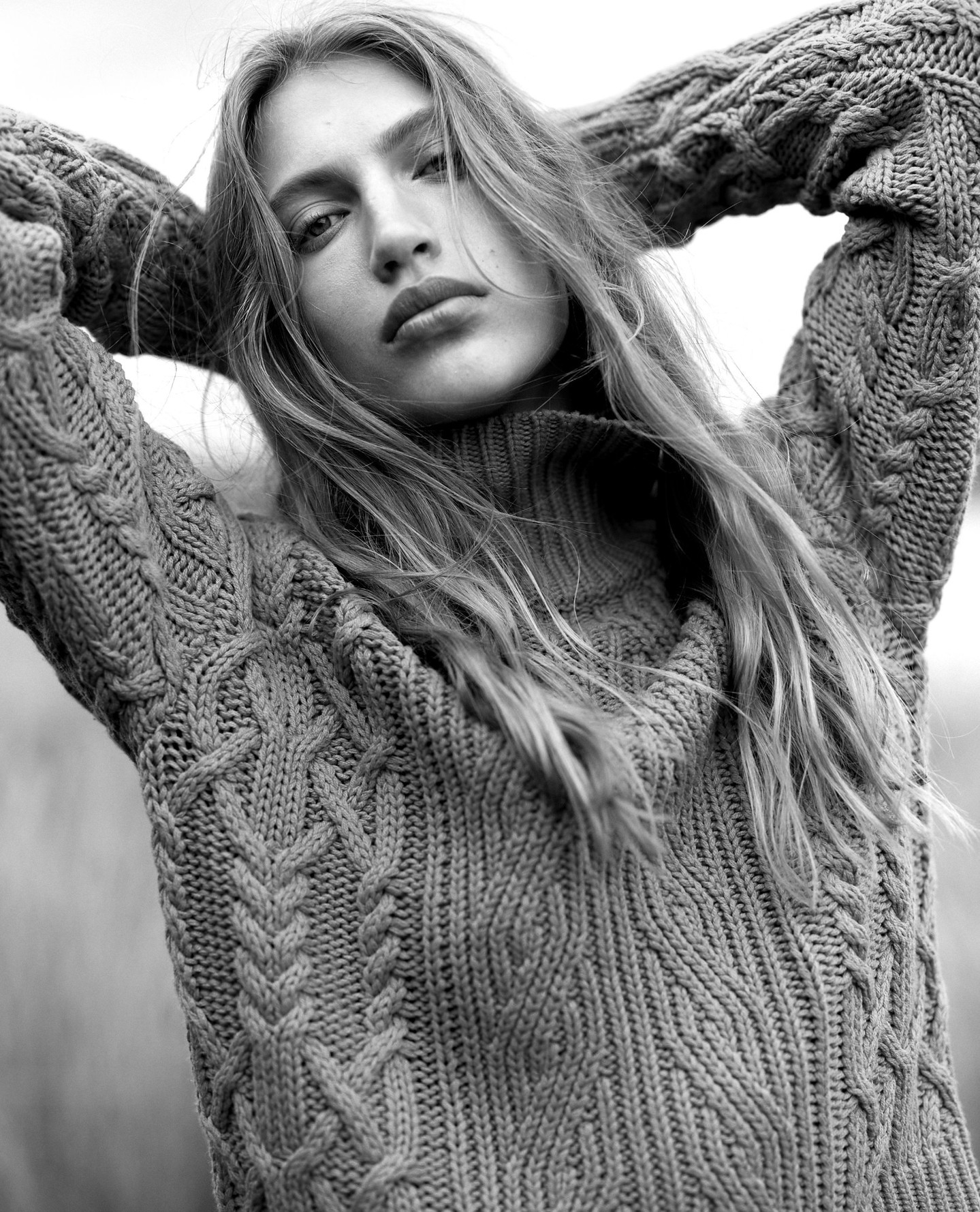 Black and white portrait of a young woman with long hair wearing a cable knit sweater, hands behind her head, outdoor setting.