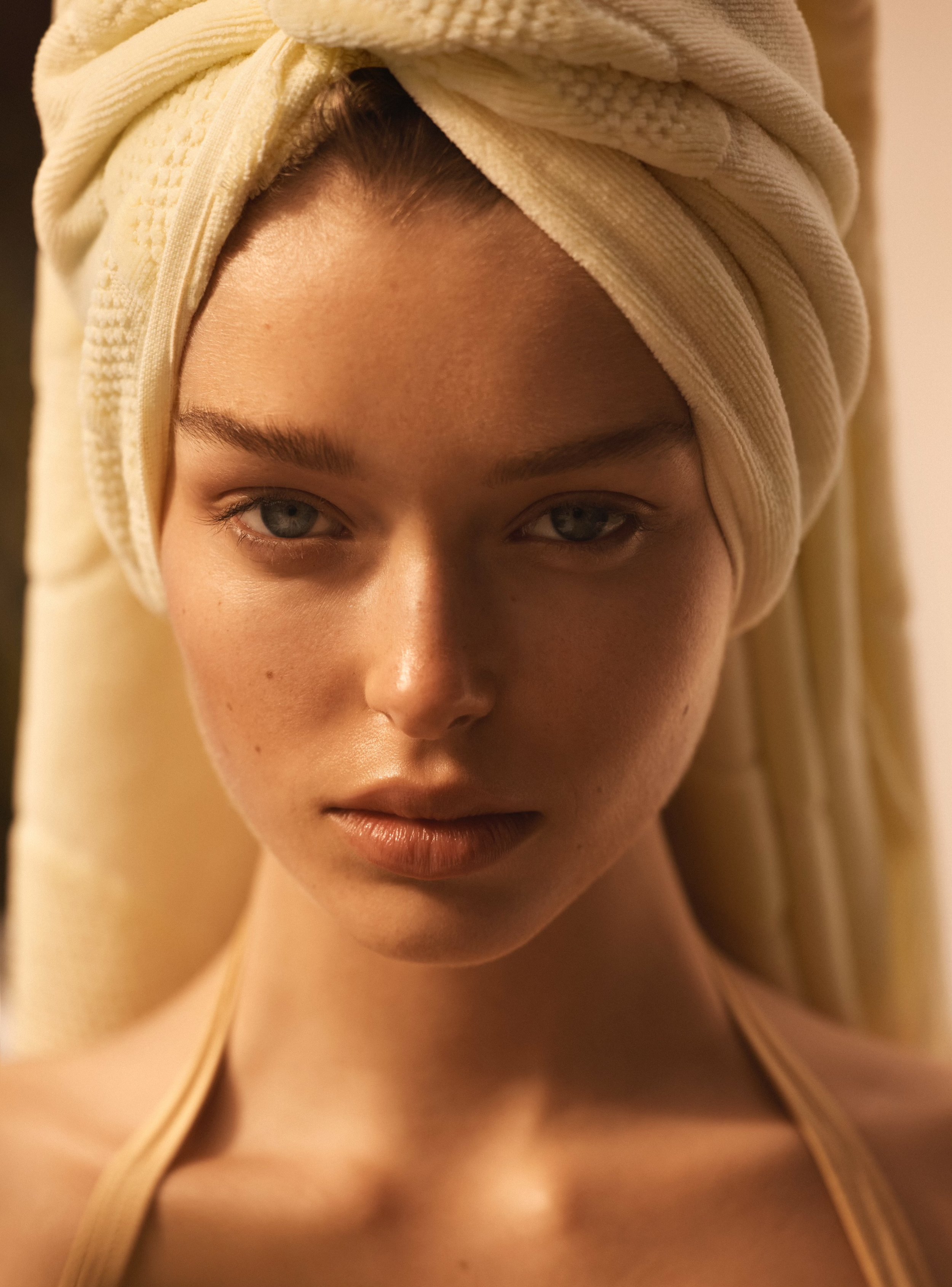 Close-up of a woman with fair skin and blue eyes, wearing a towel wrapped around her head in a turban style, soft natural lighting.