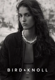 Black and white portrait of a woman with long wavy hair wearing a textured dark jacket and a shell necklace with makeup by Nadine Monley.