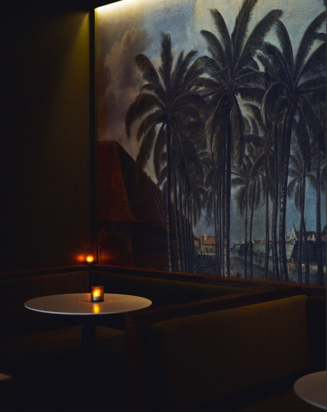 Dimly lit restaurant booth with a round white table, a small glowing candle, and a large tropical palm mural on the wall by Paul Barbera
