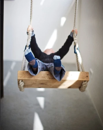 Child is swinging indoors on a wooden swing suspended by thick ropes, their back faces the camera in an eye-level shot.