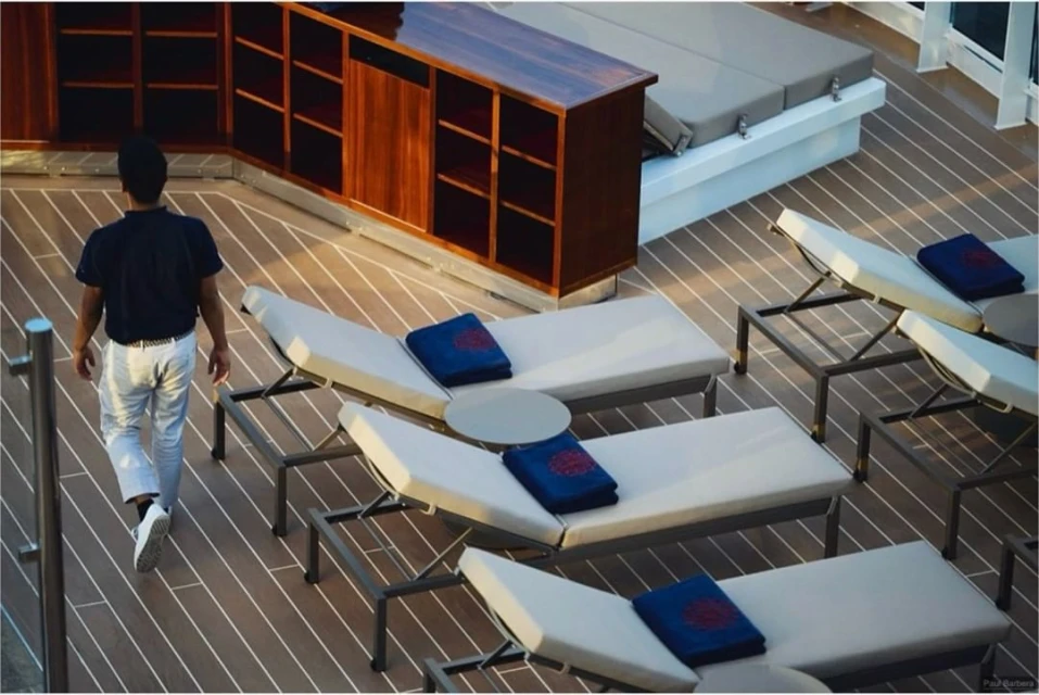 Adult male walking on the wooden deck of a cruise ship, with lounge chairs, blue towels, and dark wood storage.