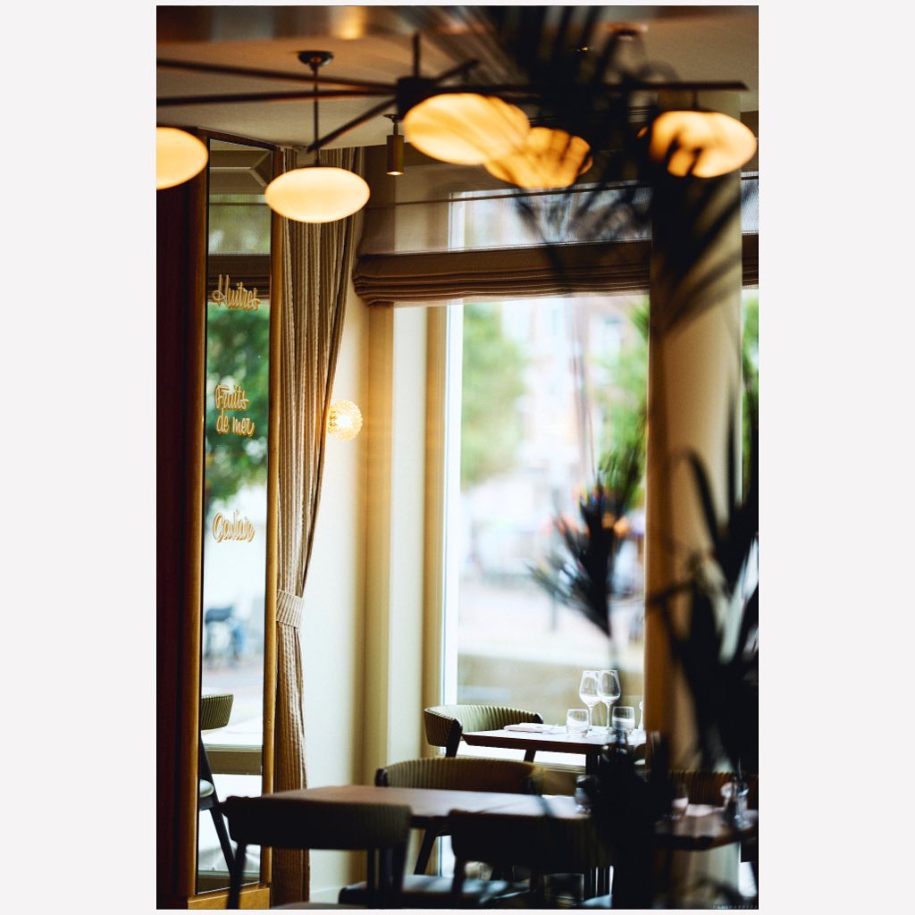 Indoor restaurant scene features tables and chairs, wine glasses, a window overlooking a city street, and natural and artificial lighting.