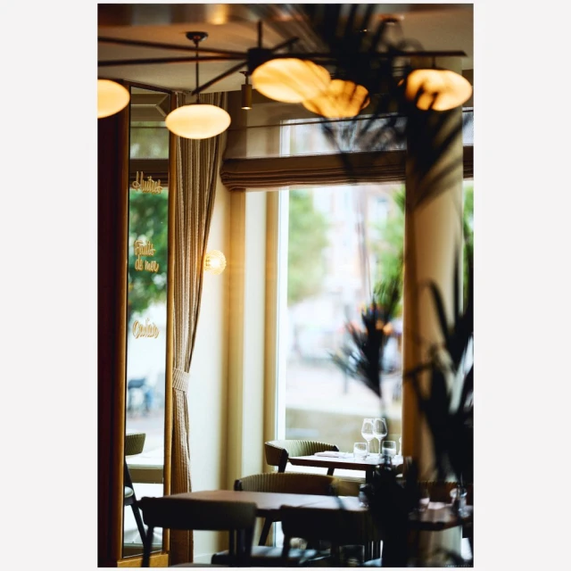 Indoor restaurant scene features tables and chairs, wine glasses, a window overlooking a city street, and natural and artificial lighting.