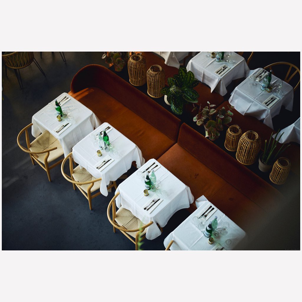 Overhead shot of multiple dining tables set with white tablecloths, silverware, bottles, and chairs, creating an inviting atmosphere.