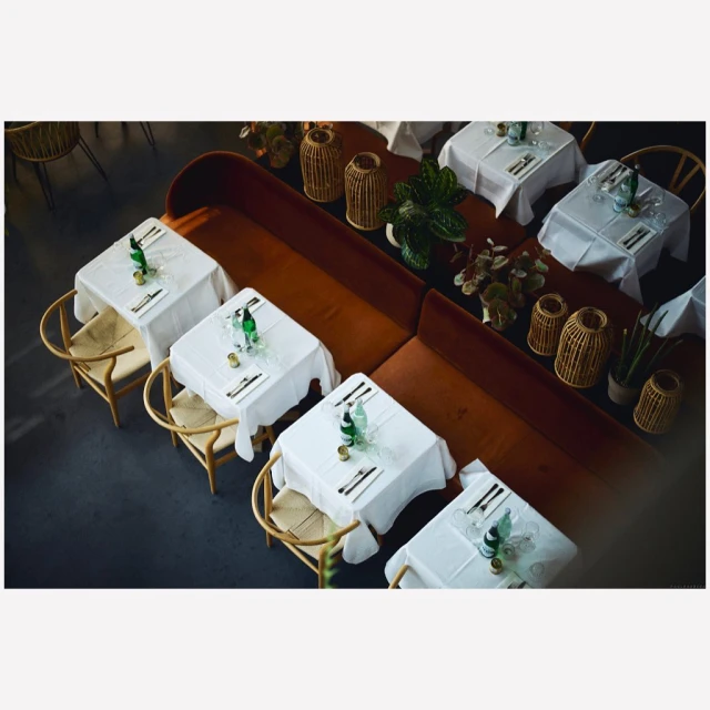 Overhead shot of multiple dining tables set with white tablecloths, silverware, bottles, and chairs, creating an inviting atmosphere.