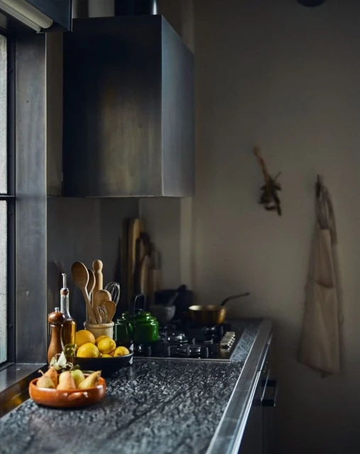Moody kitchen interior with wooden utensils, fruit bowls, and a stove under a metallic vent hood, creating a cozy culinary space.