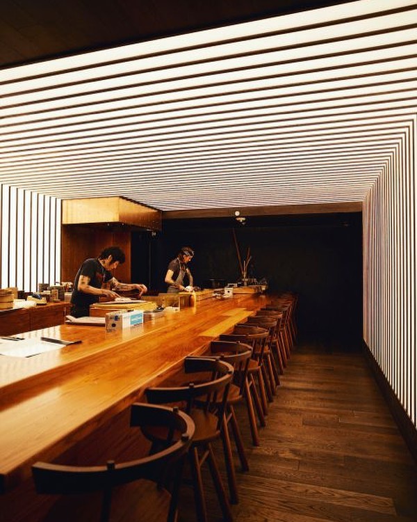 Interior of a modern restaurant featuring two chefs behind a long wooden counter with high stools, and a contemporary striped lighting design.
