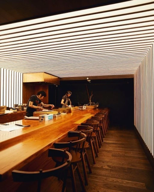 Interior of a modern restaurant featuring two chefs behind a long wooden counter with high stools, and a contemporary striped lighting design.