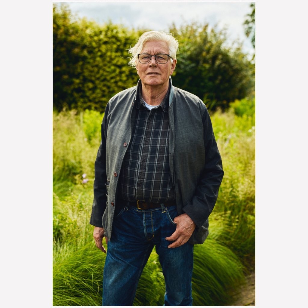 Portrait of an elderly man in a checkered shirt and jacket, standing in a green field, with a blurred background of trees and foliage.