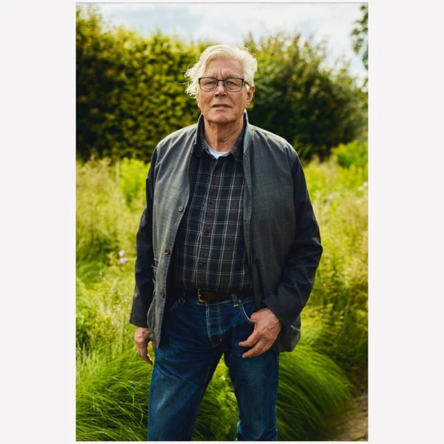 Portrait of an elderly man in a checkered shirt and jacket, standing in a green field, with a blurred background of trees and foliage.
