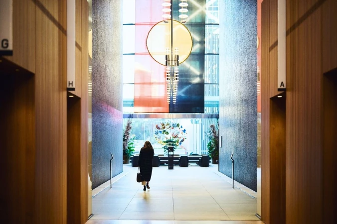 Woman walking in a modern office lobby with geometric patterns, colorful accent lighting, and wooden paneled elevator shafts. 