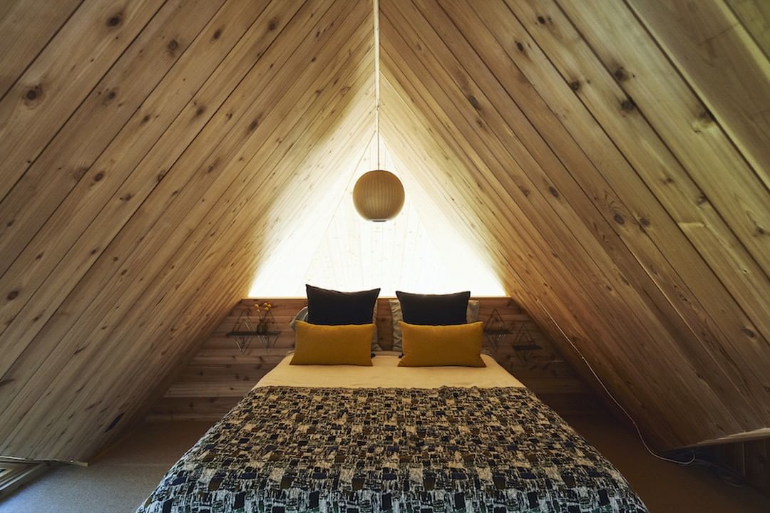 Cozy bedroom interior in an A-frame cabin, featuring a bed with patterned cover, black and yellow pillows, and wood paneled walls and ceiling.