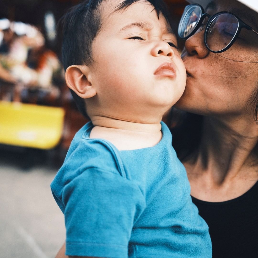 Close-up portrait of a woman wearing glasses kissing a toddler in a blue shirt on the cheek.